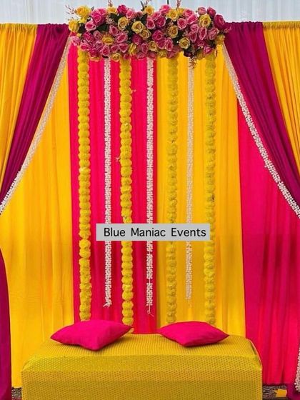A bright Haldi or Mehendi backdrop with pink and yellow drapes, accented with strings of marigolds and white flowers.