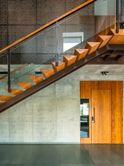 A floating wooden staircase against a raw concrete wall in J-House. The design combines industrial materials with refined craftsmanship to create a space that is both modern and warm.