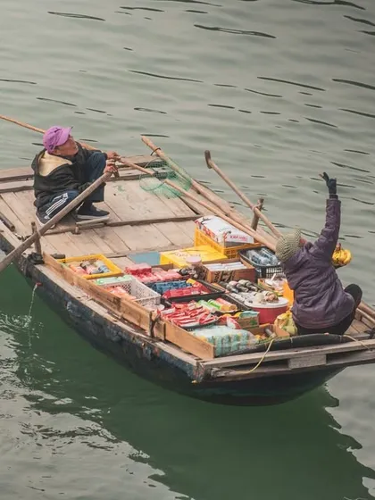 Two vendors in a small boat in Halong Bay, with one waving up towards the camera on a cruise ship.
