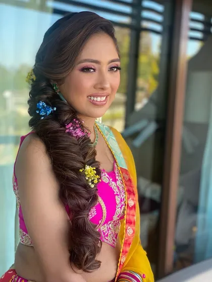Popping colors for a Mehendi look! This side-swept fishtail braid is adorned with vibrant flowers, adding a playful and colorful touch that’s perfect for the celebration.