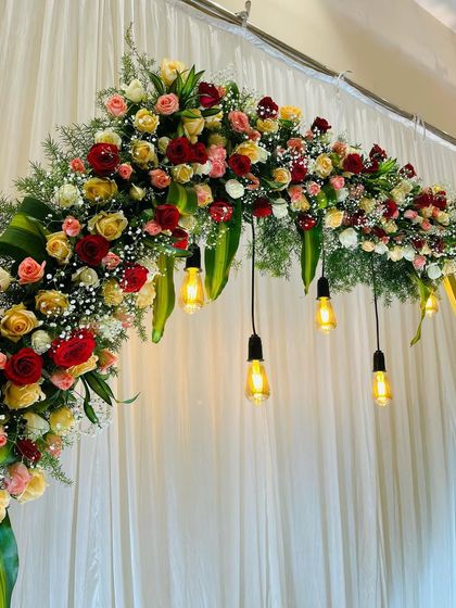 A close-up of a floral arch for an engagement stage. The arch is decorated with red and yellow roses, with hanging Edison bulbs adding a touch of modern charm.