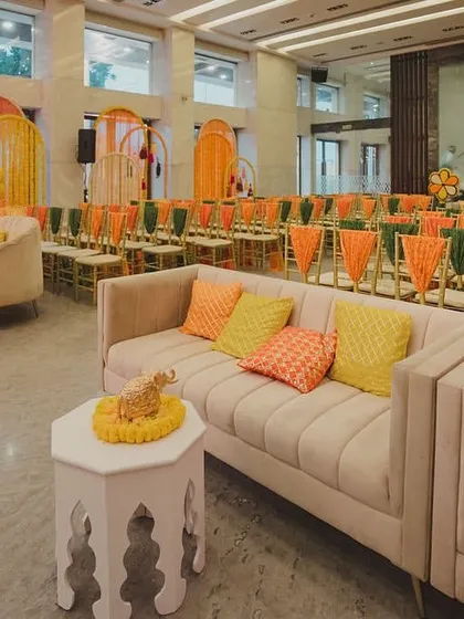 A vibrant seating area for Ayesha and Madhav's Mata ki Chowki. The decor is filled with the auspicious colors of orange and yellow, with traditional floral arrangements and comfortable lounge furniture.