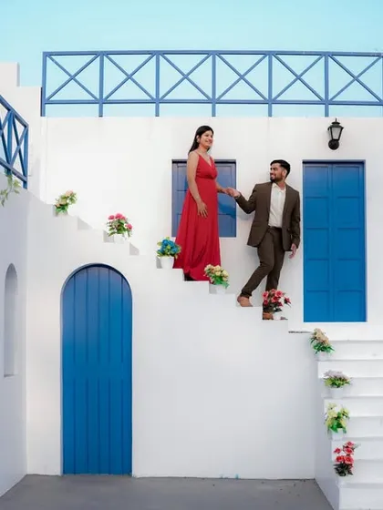 A fairytale moment on the steps of a beautiful Santorini-style building. The groom helps his bride-to-be, dressed in a flowing red gown, creating a classic and romantic cinematic shot.
