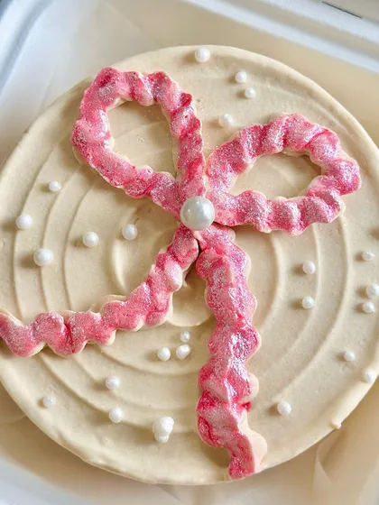 A close-up of a coquette-style bento cake, featuring a large, glittery pink bow made of buttercream and delicate pearl sprinkles.