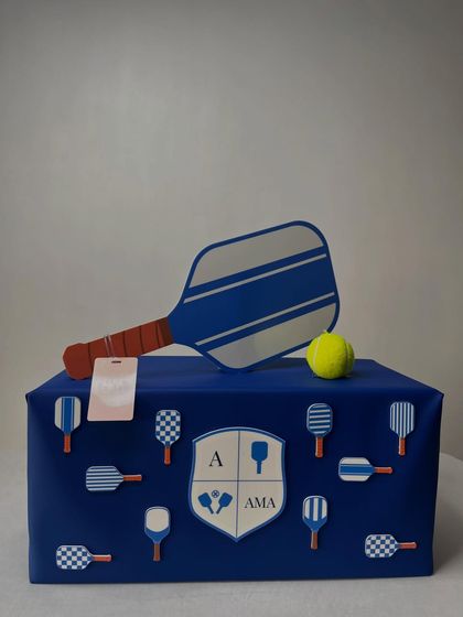 This gift celebrates a love for pickleball, featuring a custom crest with the initials 'A' and 'AMA'. The box is adorned with miniature paddle motifs and topped with a decorative paddle and ball.