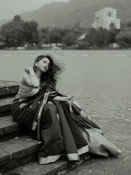 Black and white photography has a timeless power. This portrait of a woman in a saree, seated by the water with the wind in her hair, feels classic and serene. The monochrome tones emphasize the texture of the saree and the natural landscape.