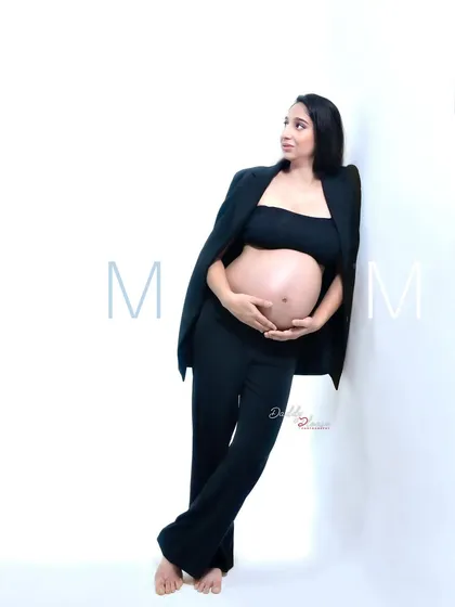 A cool and casual pose from a modern maternity session. The mom-to-be leans against the wall in her black pantsuit, creating a relaxed yet stylish portrait.