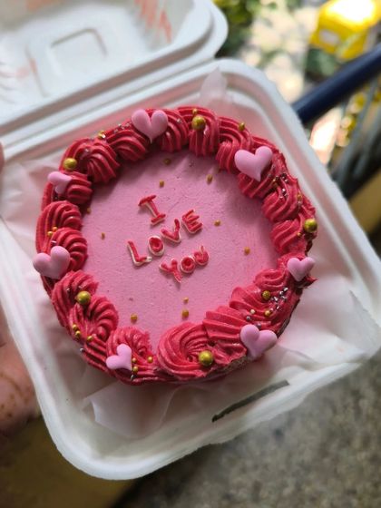 A vibrant pink bento cake that declares "I Love You." The design is framed with a detailed pink and red buttercream border and accented with gold sprinkles for an extra touch of romance.