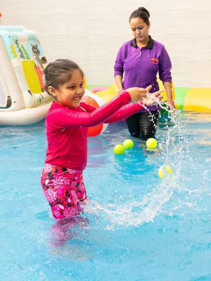 A young girl joyfully splashes water and plays with floating balls in the pool, while one of our attentive caretakers supervises nearby to ensure everyone is safe.