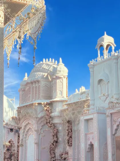 An ethereal view of a white palace structure against a brilliant blue sky. The design features delicate blush pink floral vines climbing the ornate white pillars and domes, creating a fairytale setting for a couple's special day.