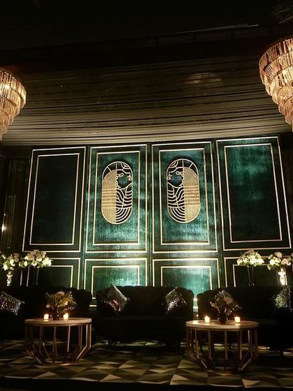 A sophisticated lounge area for a Sangeet, featuring a dark teal paneled backdrop with gold Art Deco motifs, plush velvet seating, and dramatic tiered chandeliers.