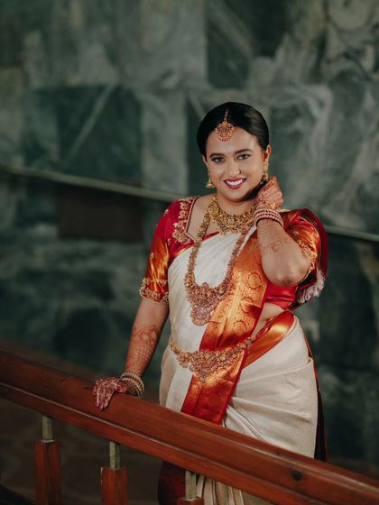 A full-length bridal portrait on a staircase, showcasing the beautiful drape of her white and red silk saree and her confident pose.