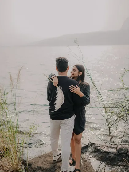 A shot from behind the couple, focusing on their embrace as they look out at the water. The feather on his back adds a unique, artistic detail to this romantic scene.