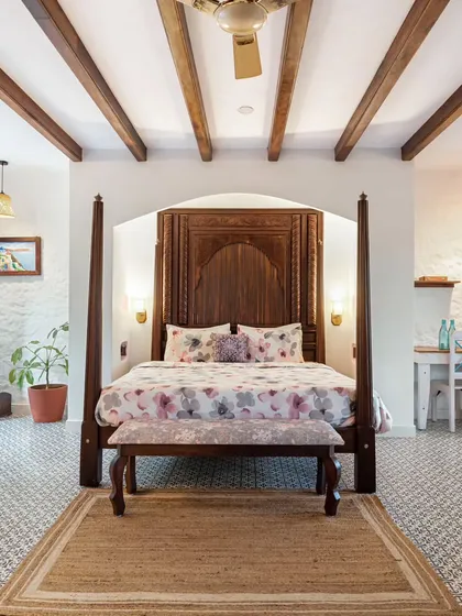 A beautiful bedroom in a Coorg villa, featuring a grand four-poster bed and exposed wooden ceiling beams. The design blends Mediterranean style with the rustic charm of a mountain retreat.