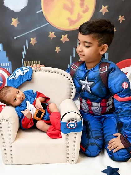 A superhero's welcome for a new baby brother. Dressed as Captain America, this big brother proudly watches over his tiny sidekick. Themed sibling photoshoots are a fun way to celebrate the start of a lifelong friendship.