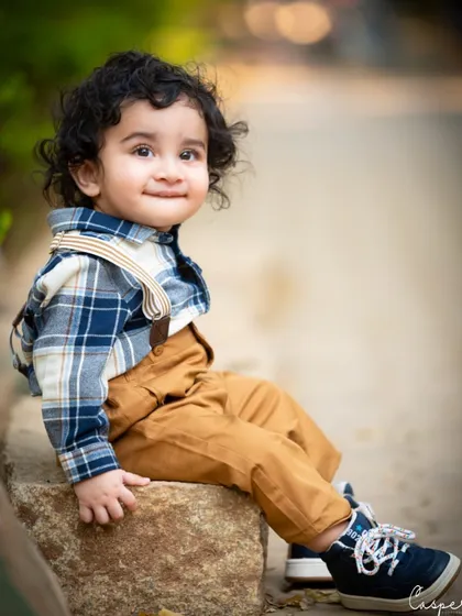 A handsome little boy with curly hair, sitting on a rock in the park. The natural setting and his stylish outfit create a timeless portrait.