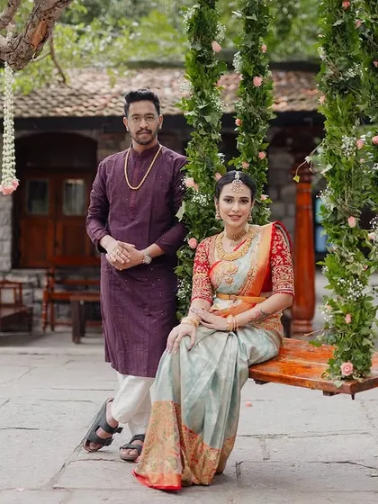 A full-length portrait of a couple by the floral swing, showcasing their complete outfits against the backdrop of the stone courtyard.
