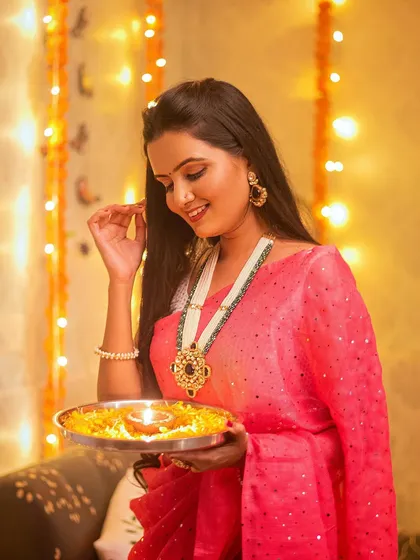 A beautiful shot of a model in a pink saree, holding a traditional puja thali. The warm lighting from the diya and fairy lights creates a magical glow.