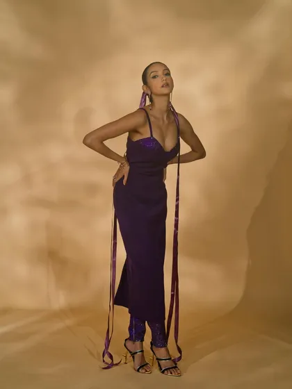 A full-length shot of a model in a purple gown with long ribbon details. The pose is dynamic, showcasing the unique design elements and modern feel of the outfit.
