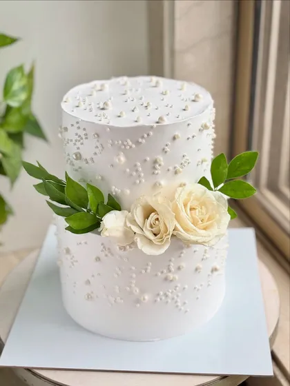 A classic two-tier wedding cake with a clean, modern aesthetic. The smooth buttercream is decorated with scattered pearls and a simple arrangement of fresh white roses and green leaves.