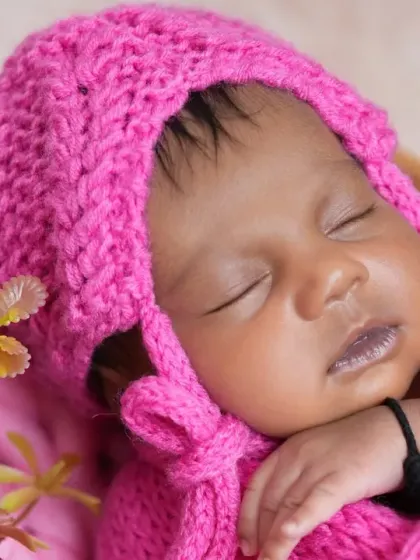 A close-up shot of a newborn's face, snugly wrapped in a pink knit bonnet and swaddle. The focus is on the baby's peaceful expression and tiny, delicate features.
