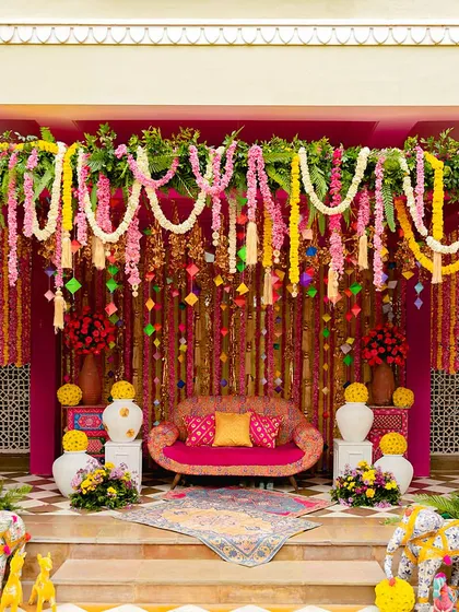 A vibrant and traditional bhaat ceremony. The seating area is a riot of pink and yellow, with floral garlands, colorful elephants, and a beautifully decorated sofa.