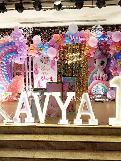 A magical unicorn and rainbow themed first birthday stage for Kavya. The setup includes a large rainbow balloon arch, a cute unicorn prop, a sparkling gold sequin backdrop, and illuminated marquee letters, creating a truly enchanting scene.