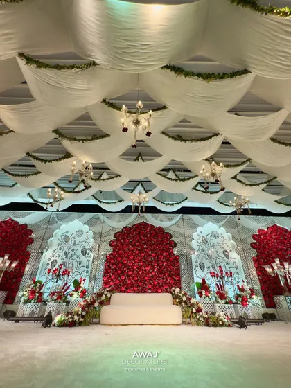 A regal reception stage where a canopy of white fabric and greenery drapes over a stunning backdrop of red roses, creating a look of classic opulence and romance.