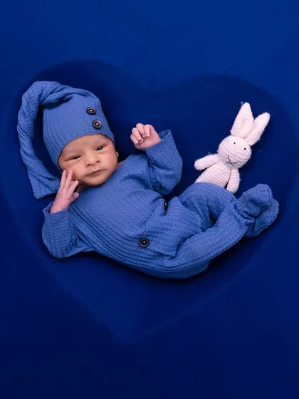 A newborn boy in a blue outfit rests inside a heart-shaped prop, accompanied by a small crochet bunny.