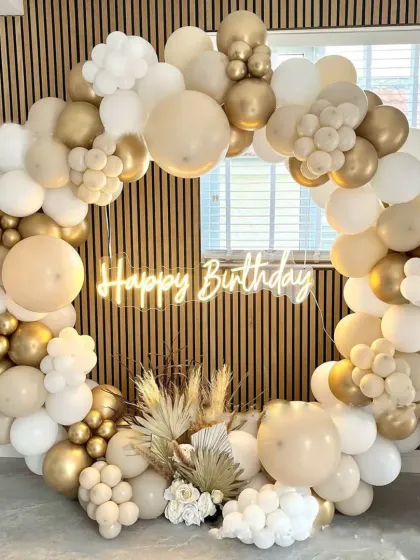 If you want an elegant and modern look, I can do that too. This birthday setup features a stylish balloon ring using white, beige, and gold chrome balloons, complete with a neon sign and dried pampas grass for a sophisticated touch.