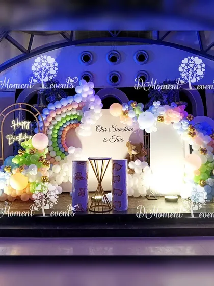 A beautiful rainbow and teddy bear themed stage for a second birthday, with a large balloon rainbow and "Our Sunshine is Two" message.