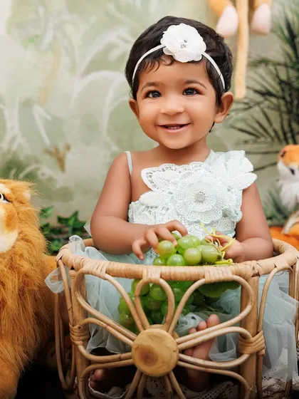 A smiling baby girl holds a bunch of grapes while sitting in a basket, surrounded by her friendly jungle animal toys.