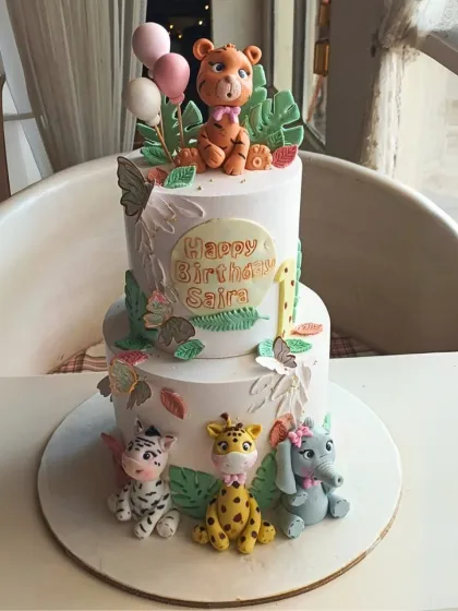An adorable two-tier jungle safari cake featuring cute, handcrafted fondant animals like a tiger, zebra, giraffe, and elephant for a first birthday.