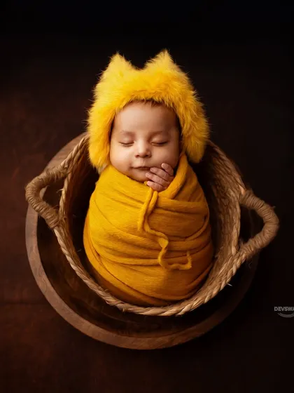 A classic and adorable posed newborn photo. The baby is comfortably swaddled in a vibrant yellow wrap and a cute, furry hat, nestled safely in a woven basket for a perfect, sleepy portrait.