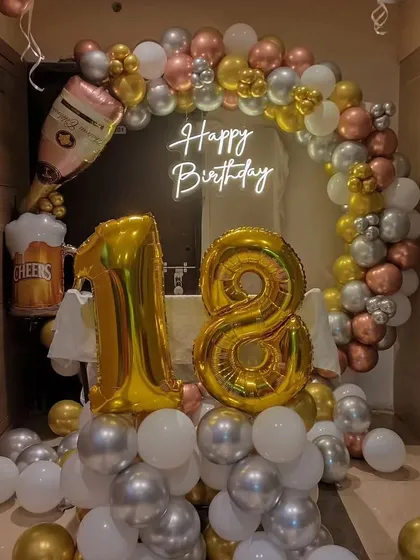 A fun and festive 18th birthday decoration. The setup includes a balloon arch with champagne and beer foil balloons, a neon sign, and large gold foil numbers.