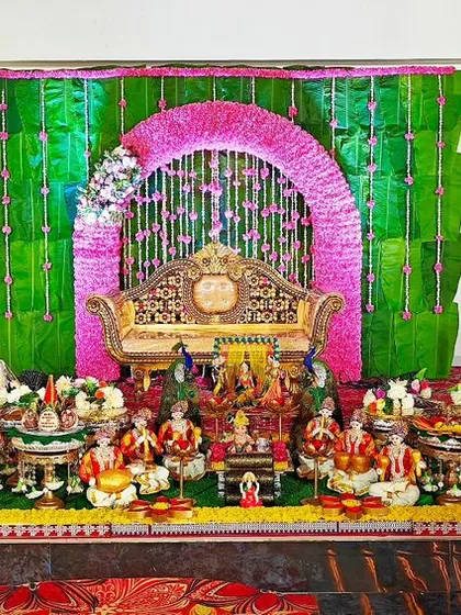 This baby shower setup features a vibrant green and pink floral arch over a traditional seat. The display is filled with dolls representing family and celebration, making it a lively and joyous scene.