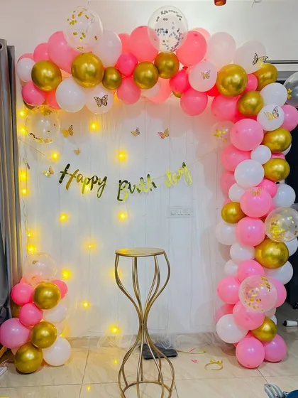 A delicate and pretty birthday setup with a pink, white, and gold balloon arch, fairy lights, and butterfly accents.