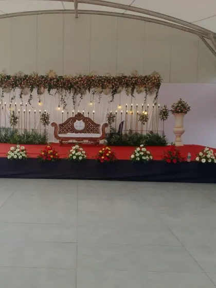 This is a full view of an elegant and simple stage design. It features a classic carved sofa for the couple, a backdrop of white drapes with hanging lights, and floral arrangements in large urns, all set on a red carpeted stage.