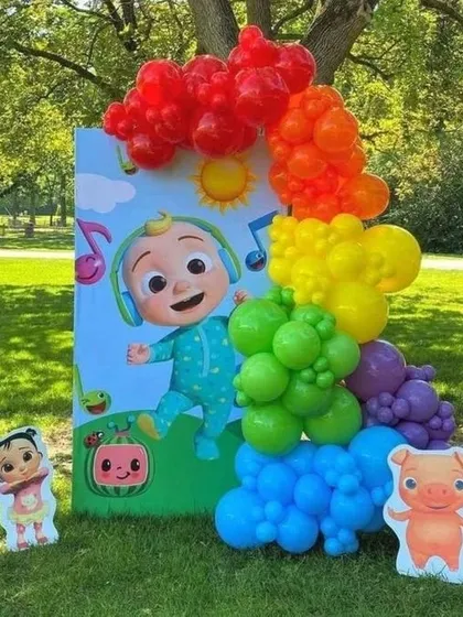 An outdoor Cocomelon themed decoration, perfect for a backyard birthday party. The setup includes a large printed backdrop with the main character, a rainbow balloon garland, and smaller cutouts of other characters from the show.