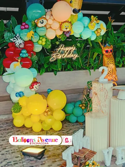 A close-up of a jungle-themed birthday decoration. This setup includes a neon "Happy Birthday" sign, animal foil balloons, and a colorful balloon garland against a natural green background.