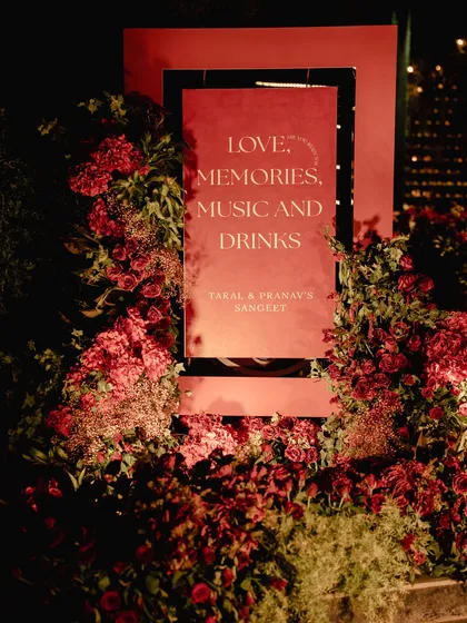 The welcome signage for the red Sangeet. A mirrored sign with the words "Love, Memories, Music and Drinks" is nestled within a bed of lush red flowers, setting the tone for the night.