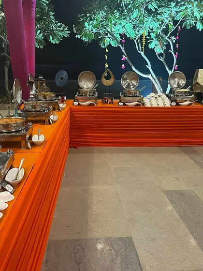 A vibrant and colourful buffet setup for a Mehandi ceremony. The bright orange table linens and decorative trees create a festive atmosphere perfect for pre-wedding celebrations.