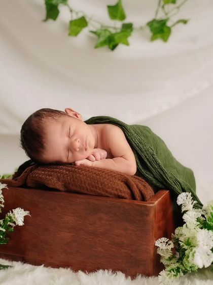 A newborn sleeps on their side on a wooden box, partially covered by a green wrap and surrounded by white flowers.