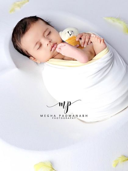 A wider view of the baby and his teddy bear, nestled in a heart-shaped prop with soft yellow feathers.