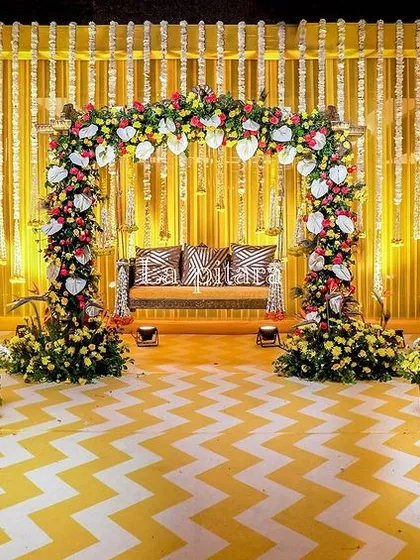 A stunning stage setup for a traditional Teej celebration. The design features a vibrant yellow backdrop, a floral-draped swing, and magnificent peacocks crafted entirely from fresh white flowers, blending cultural symbolism with artistic floral design for a truly memorable event.