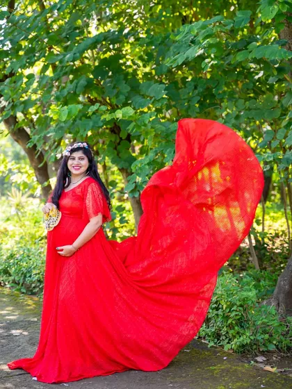 A vibrant shot of the mom-to-be in a flowing red lace gown, standing under a canopy of green leaves.