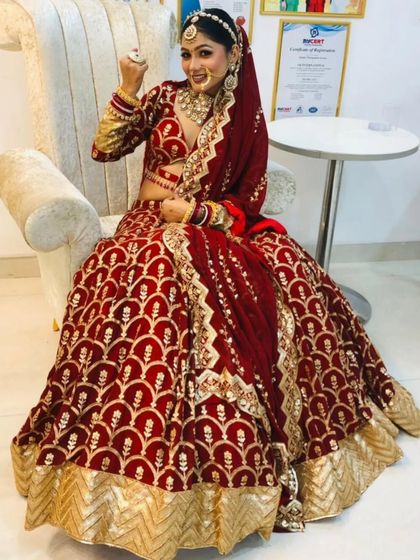 A happy bride seated in a chair, showing the full flare of the maroon arched-pattern lehenga.