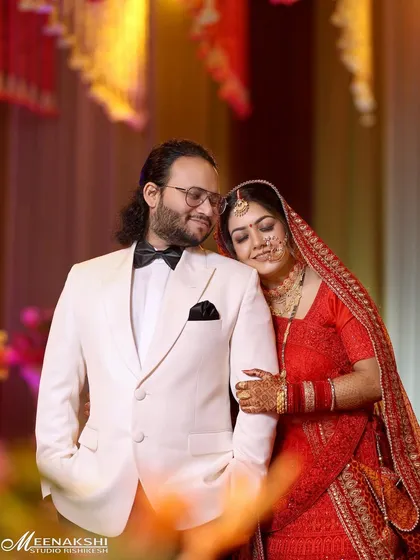 A classic wedding portrait of the couple. The bride in her traditional red lehenga rests her head on the groom's shoulder, who is dressed in a sharp white tuxedo, creating a beautiful contrast.