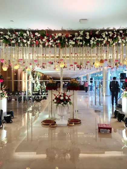 A grand and opulent welcome aisle decorated with hanging floral arrangements and traditional brass platters. The polished marble floor reflects the lights and flowers, doubling the beauty.
