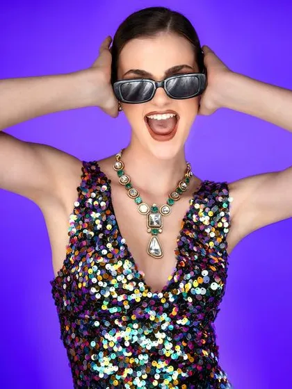Fun, energetic, and full of life. The combination of a sequined dress, sunglasses, and a joyful expression against a solid purple background creates a dynamic and playful party look.